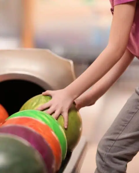 A person wearing a pink shirt and gray pants selects a green bowling ball from a rack of colorful bowling balls at a kids bowl free event at the bowling alley.