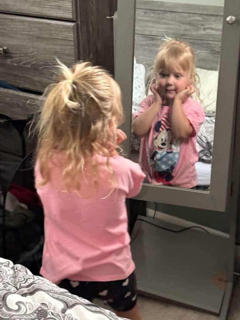 A young girl with blonde hair looks at herself in a mirror, wearing sticker earrings, a pink Minnie Mouse shirt, and black shorts with white hearts.