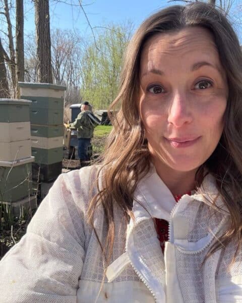 A woman in a beekeeping suit takes a selfie outdoors near beehives during a sweet spring day; another person in a suit works among the trees in the background.