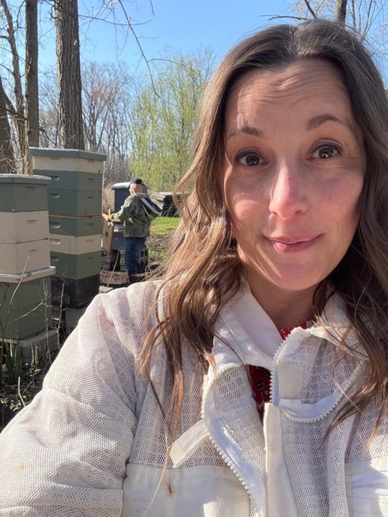 A woman in a beekeeping suit takes a selfie outdoors near beehives during a sweet spring day; another person in a suit works among the trees in the background.