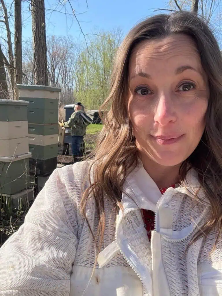 A woman in a beekeeping suit takes a selfie outdoors near beehives during a sweet spring day; another person in a suit works among the trees in the background.
