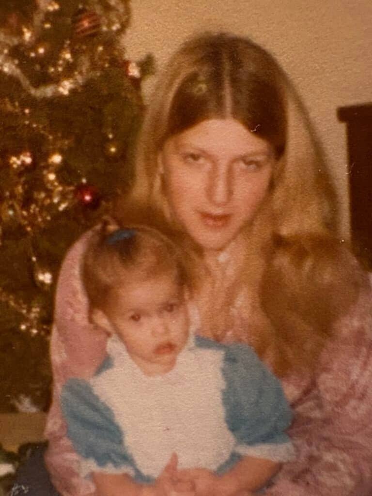 A woman with long blonde hair holds a young girl in a blue and white dress, celebrating tradition as they sit together in front of a decorated Christmas tree.