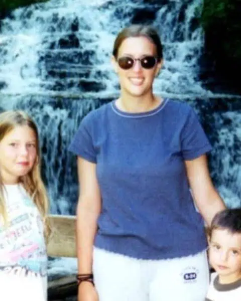 A woman and two children, representing family across generations, stand in front of a wooden railing with a waterfall and lush greenery in the background.