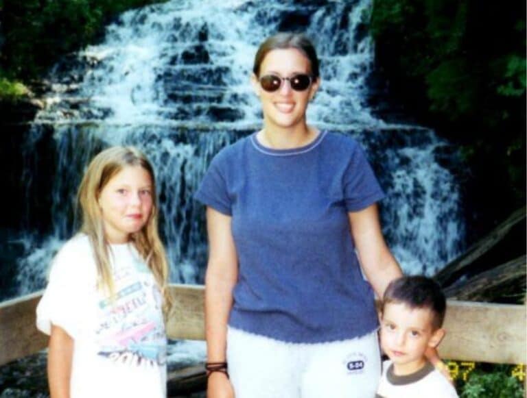 A woman and two children, representing family across generations, stand in front of a wooden railing with a waterfall and lush greenery in the background.