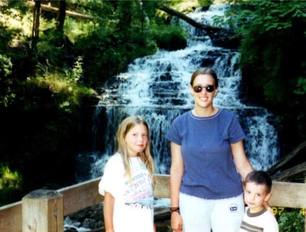 A woman and two children, representing the legacy of a family farm, stand in front of a wooden railing with a waterfall and lush greenery in the background.