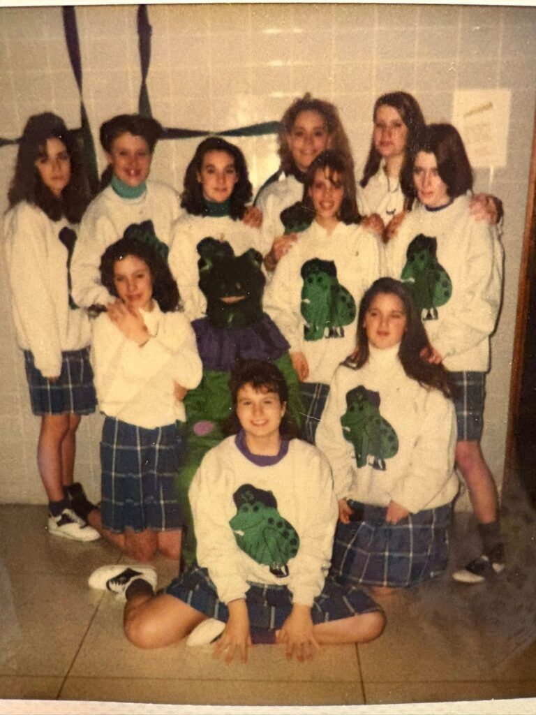 A group of girls in plaid skirts and matching white sweatshirts with green frog designs pose together indoors against a tiled wall for an update post.