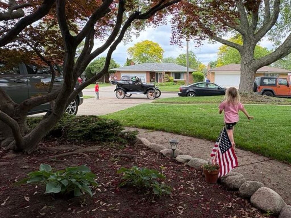 A girl holding an American flag walks on a lawn, sporting sticker earrings, while a vintage car drives by and another child stands nearby in a suburban neighborhood.