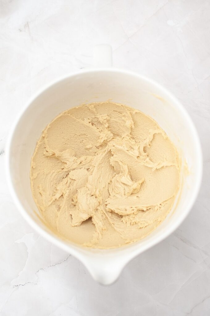 A white mixing bowl containing creamy, light brown homemade cookies dough on a white marble surface.