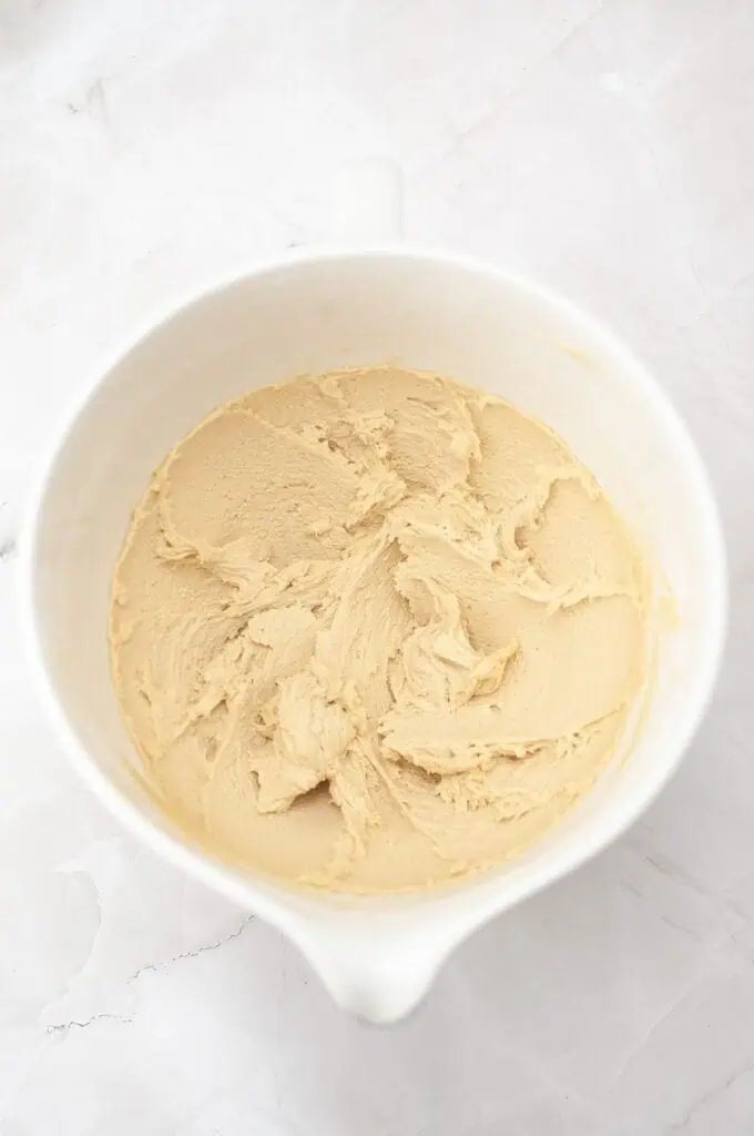 A white mixing bowl containing creamy, light brown homemade cookies dough on a white marble surface.