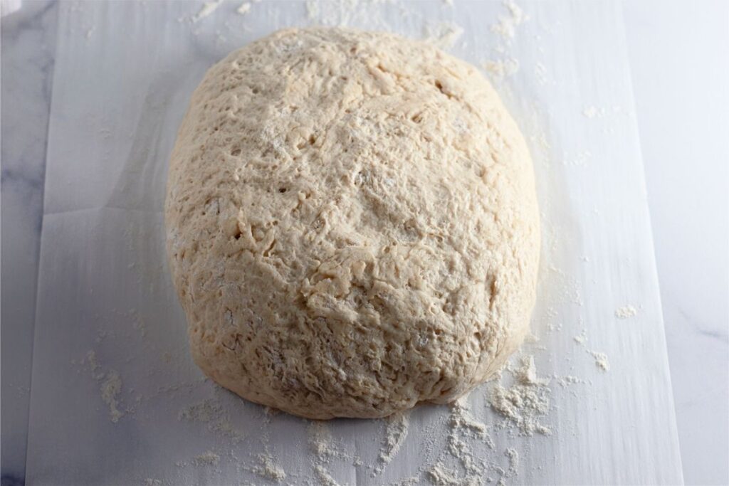 A ball of raw dutch oven bread dough rests on a lightly floured white surface.
