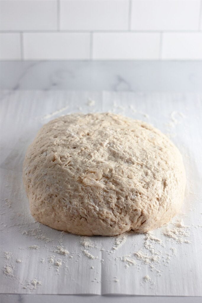 A round ball of bread dough, perfect for making Dutch oven bread, rests on a floured surface with a white tile background.
