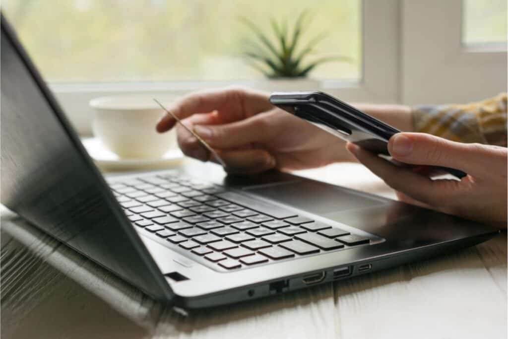 A person holds a credit card and smartphone while using a laptop on a table near a window, researching tips for negotiating lower bills with service providers; a cup and small plant sit in the background.