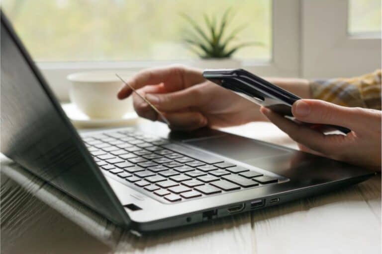 A person holds a credit card and smartphone while using a laptop on a table near a window, researching tips for negotiating lower bills with service providers; a cup and small plant sit in the background.