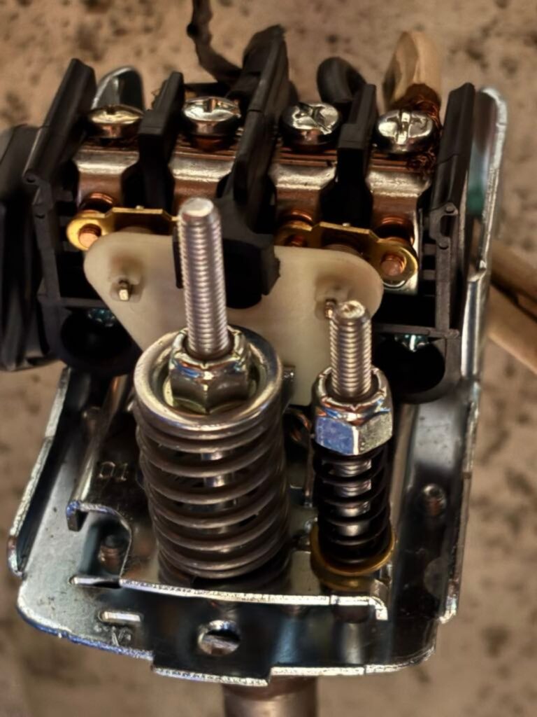 Close-up of an electrical component with exposed wires, screws, a sweet spring, and bolts mounted on a metal base.