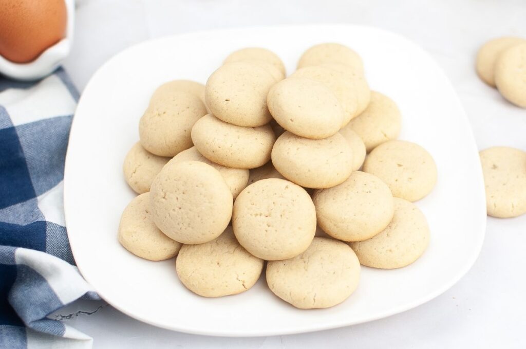 A white plate holds a pile of round, light brown homemade vanilla wafers. A blue and white napkin and a brown egg are partially visible on the side, hinting at the classic cookies recipe.