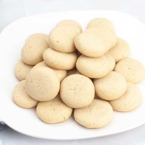 A white plate holds a pile of round, light brown homemade vanilla wafers. A blue and white napkin and a brown egg are partially visible on the side, hinting at the classic cookies recipe.