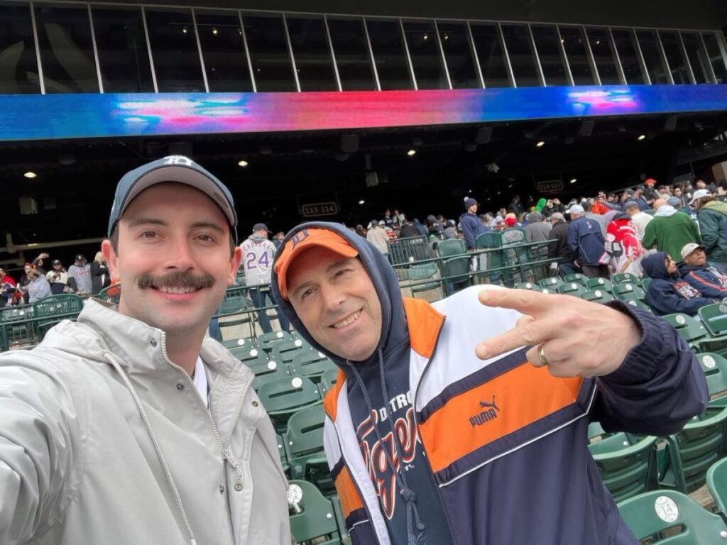 Two men pose for a selfie at a baseball stadium during a spring update; one smiles as the other flashes a peace sign, with crowds filling the seats in the background.
