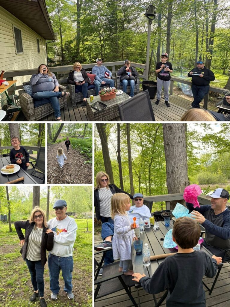 A family gathers on a wooden deck and in a yard, talking, eating stadium dogs, and playing with children wearing sticker earrings on a sunny day surrounded by trees.