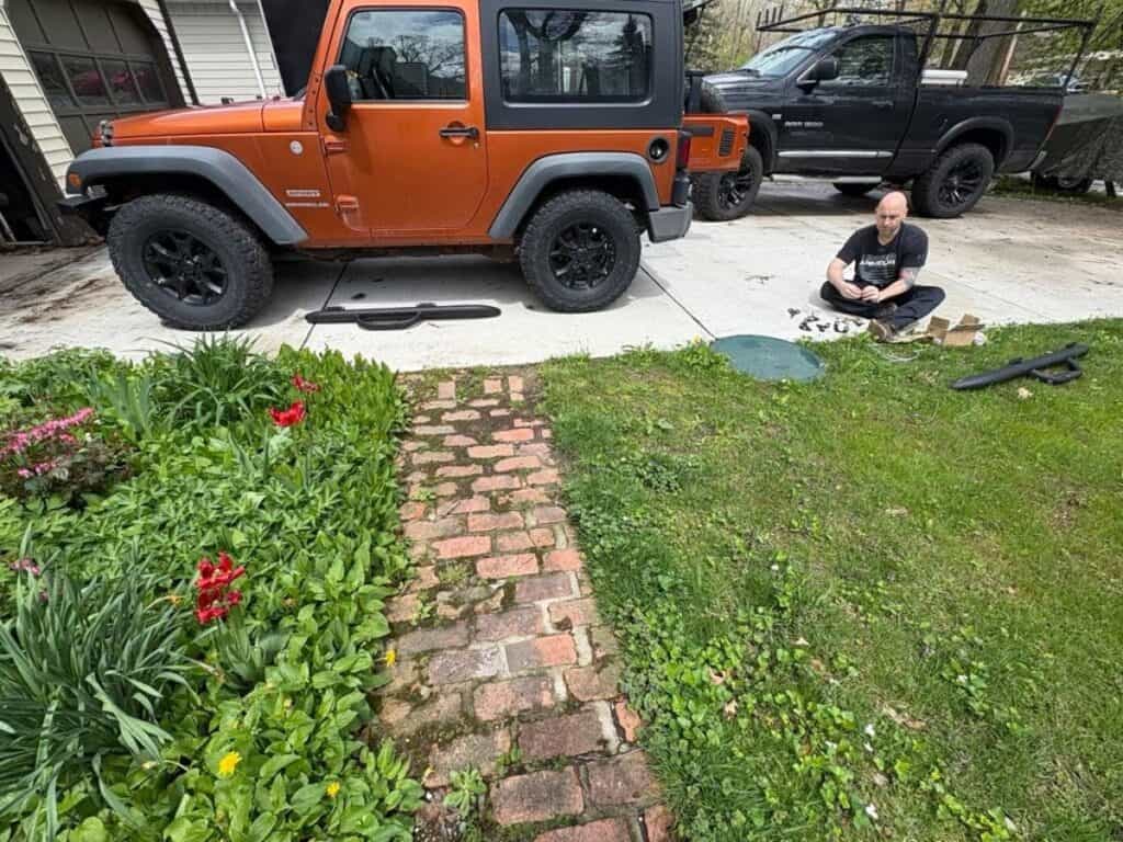 A person sits on a driveway in this personal post, assembling parts next to an orange Jeep Wrangler, with a black truck parked behind and a garden with flowers blooming in the foreground.