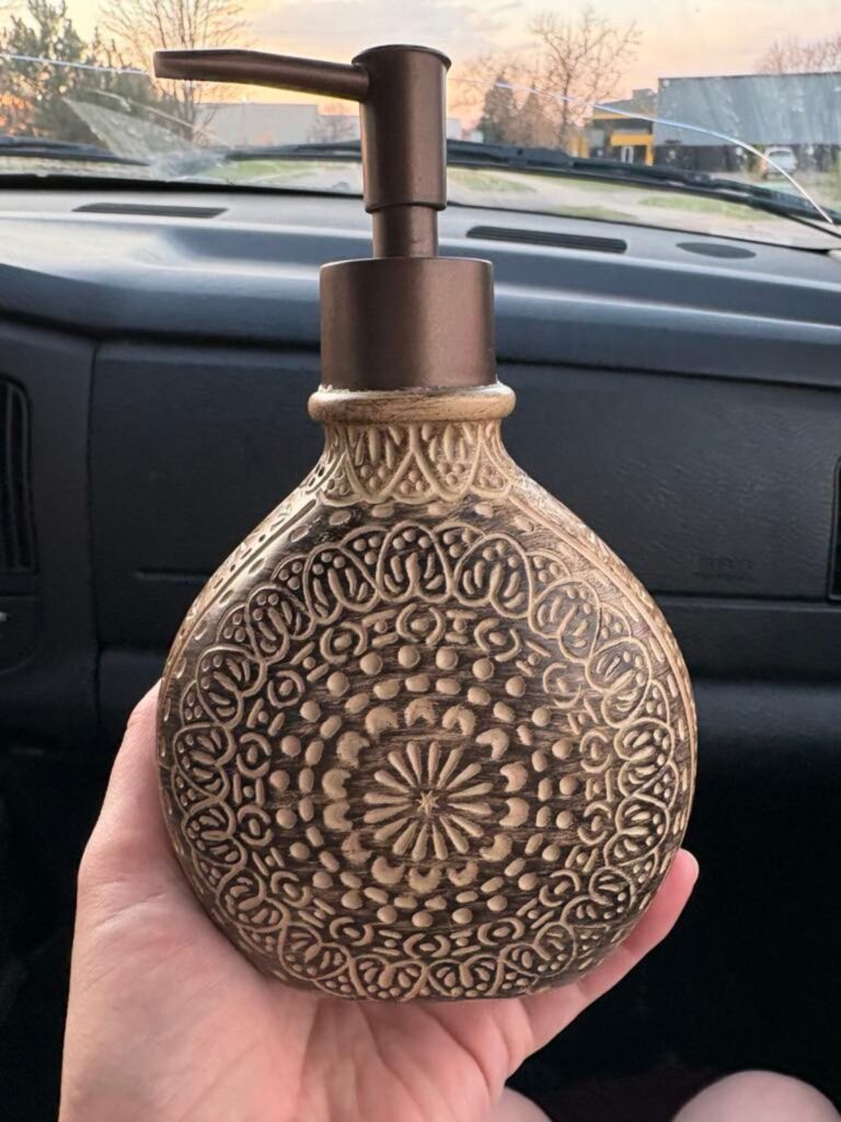 A hand holds a sweet, decorative patterned soap dispenser with a bronze pump inside a car; trees and houses hint at a Spring update through the windshield.