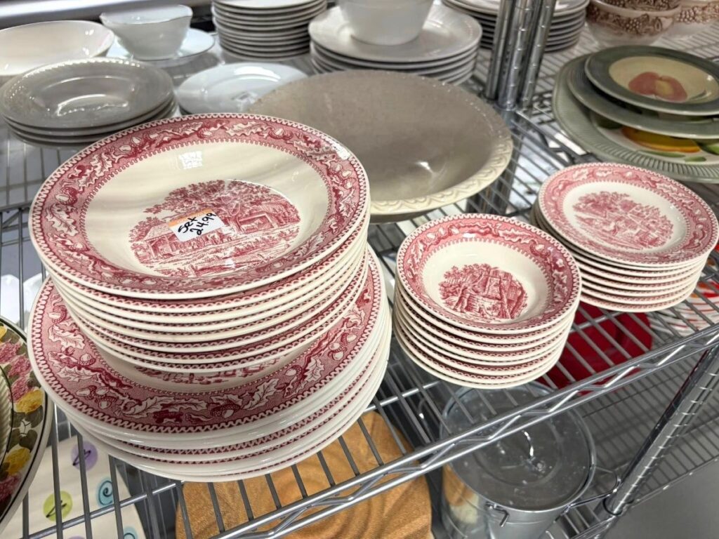 Sets of vintage red and white patterned dishes, including plates and bowls, are stacked on metal shelving in a store. A price sticker is visible on one of the plates.