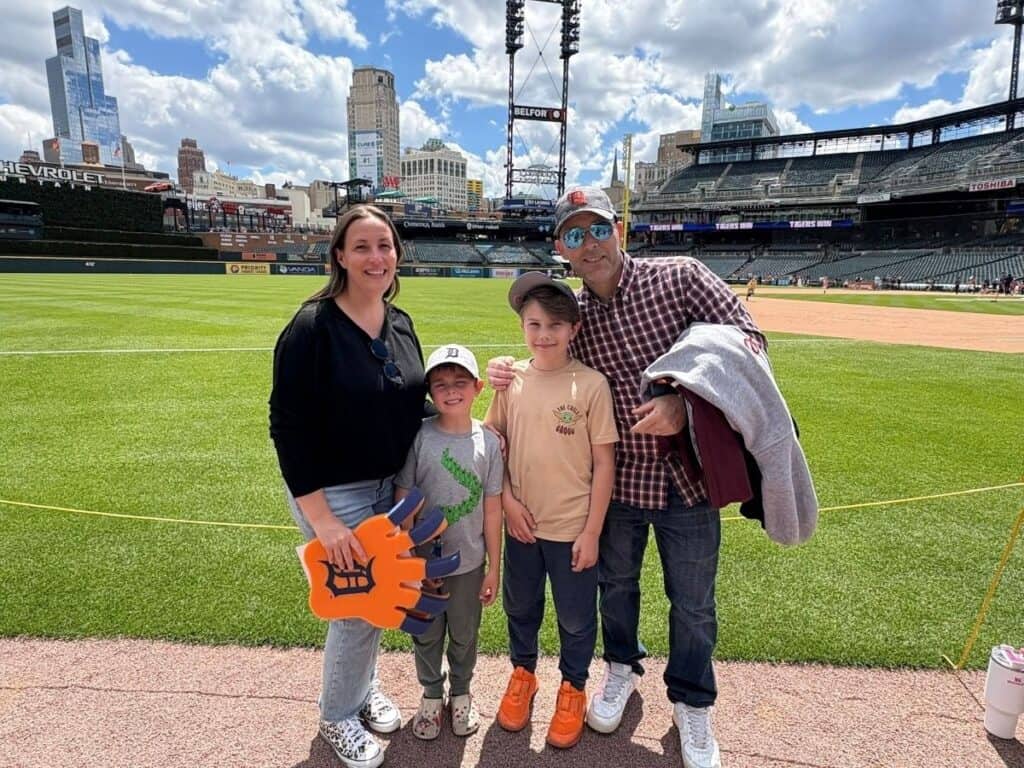 A family of four stands on the edge of a baseball field with empty stadium seats in the background. One child holds a large foam paw, while everyone beams at the camera, ready to cheer for the Stadium Dogs.