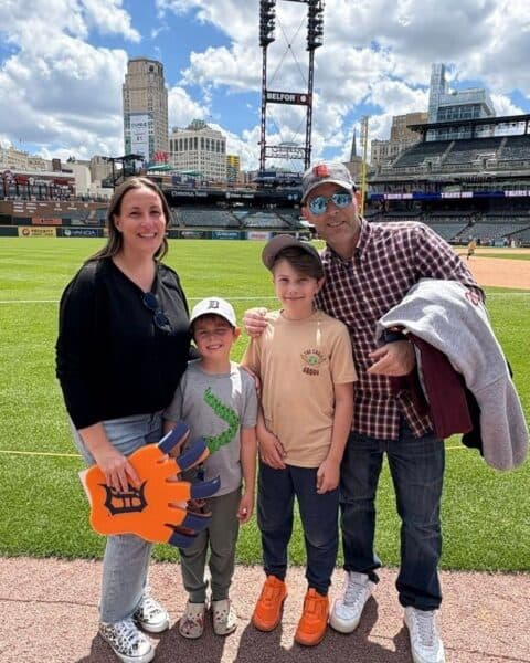 A family of four stands on the edge of a baseball field with empty stadium seats in the background. One child holds a large foam paw, while everyone beams at the camera, ready to cheer for the Stadium Dogs.