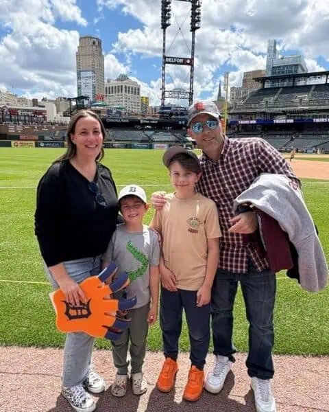A family of four stands on the edge of a baseball field with empty stadium seats in the background. One child holds a large foam paw, while everyone beams at the camera, ready to cheer for the Stadium Dogs.