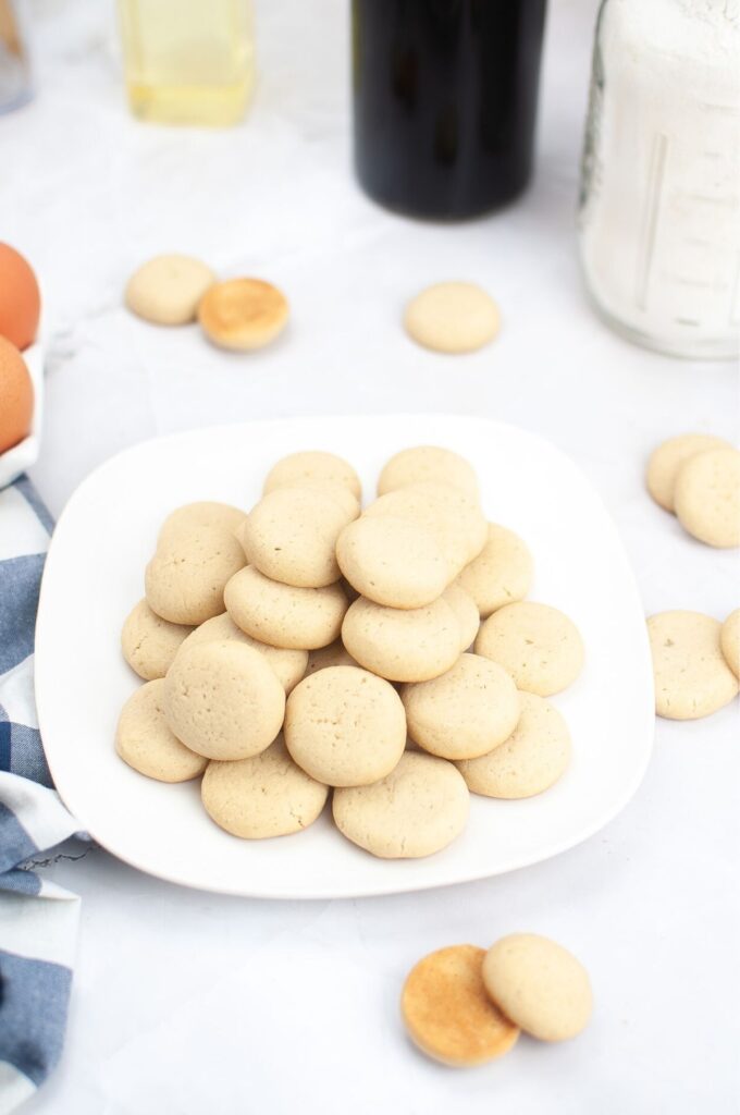 A white plate stacked with plain round vanilla cookies sits on a white surface, surrounded by a few scattered cookies and baking ingredients—perfect for trying your favorite homemade cookies recipe.