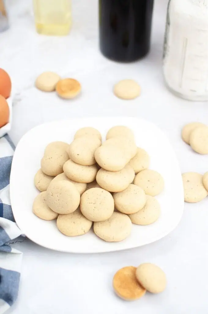 A white plate stacked with plain round vanilla cookies sits on a white surface, surrounded by a few scattered cookies and baking ingredients—perfect for trying your favorite homemade cookies recipe.