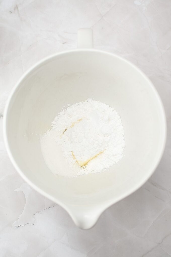 A white mixing bowl containing a block of butter, powdered sugar, and granulated sugar sits on a light marble surface—the perfect start for a classic homemade cookies recipe.