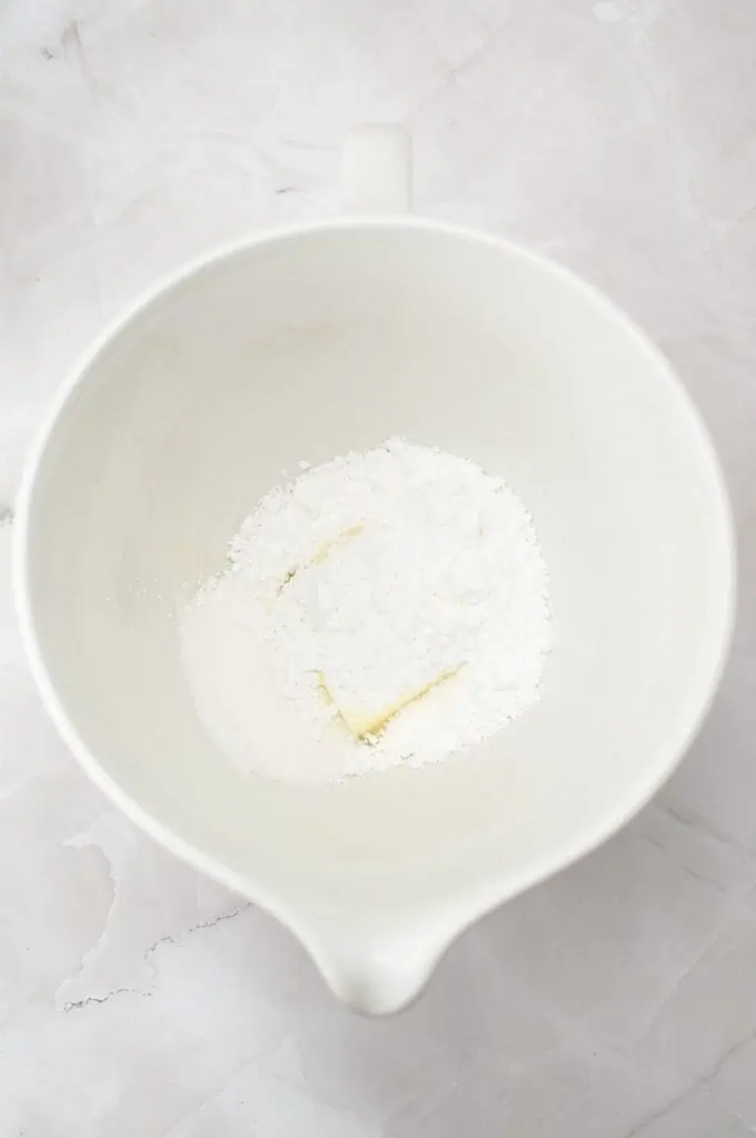 A white mixing bowl containing a block of butter, powdered sugar, and granulated sugar sits on a light marble surface—the perfect start for a classic homemade cookies recipe.