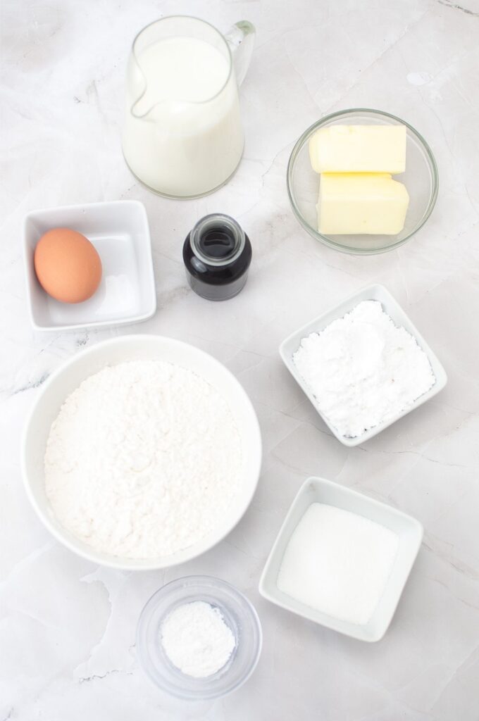 Baking ingredients on a white surface, perfect for homemade cookies or your favorite cookies recipe, including flour, sugar, butter, an egg, milk, vanilla extract, baking powder, and powdered sugar in small bowls and containers.