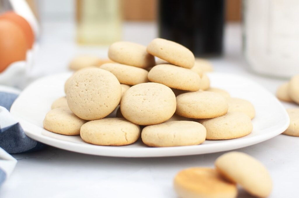 A white plate stacked with round, pale vanilla wafers sits on a light surface, hinting at a delicious homemade cookies recipe amid blurred kitchen items in the background.