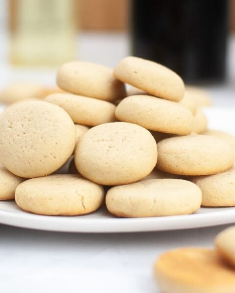 A white plate stacked with round, pale vanilla wafers sits on a light surface, hinting at a delicious homemade cookies recipe amid blurred kitchen items in the background.