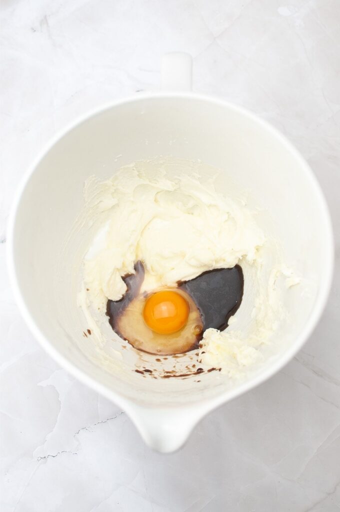 A mixing bowl containing softened cream cheese, an egg, and vanilla extract on a white marble surface—perfect beginnings for a homemade vanilla wafers recipe.
