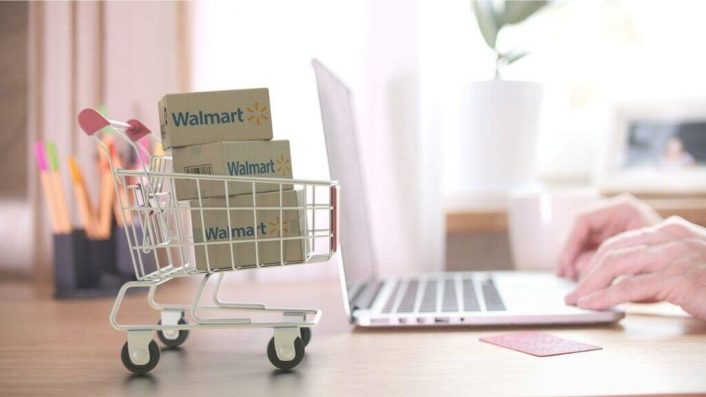 A person uses a laptop near a small shopping cart filled with Walmart boxes and a credit card on the desk, browsing to discover the best Walmart deals and new ways to save money at Walmart.