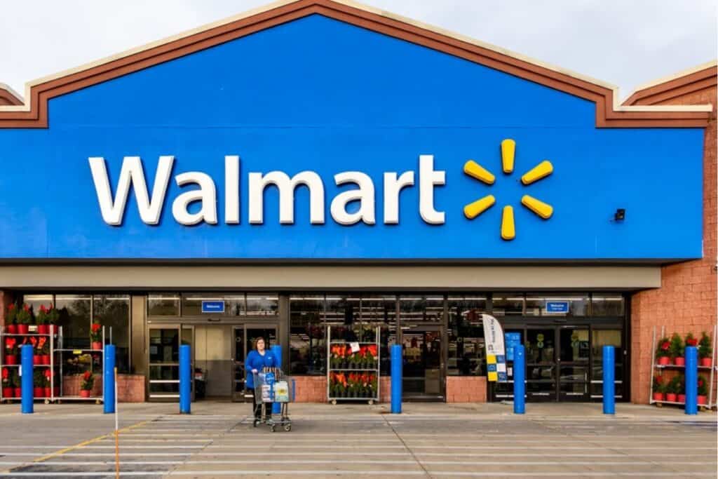 The front entrance of a Walmart store with a person pushing a shopping cart in the parking lot, visible under a blue Walmart sign, highlighting ways to Save Money at Walmart on everyday essentials.