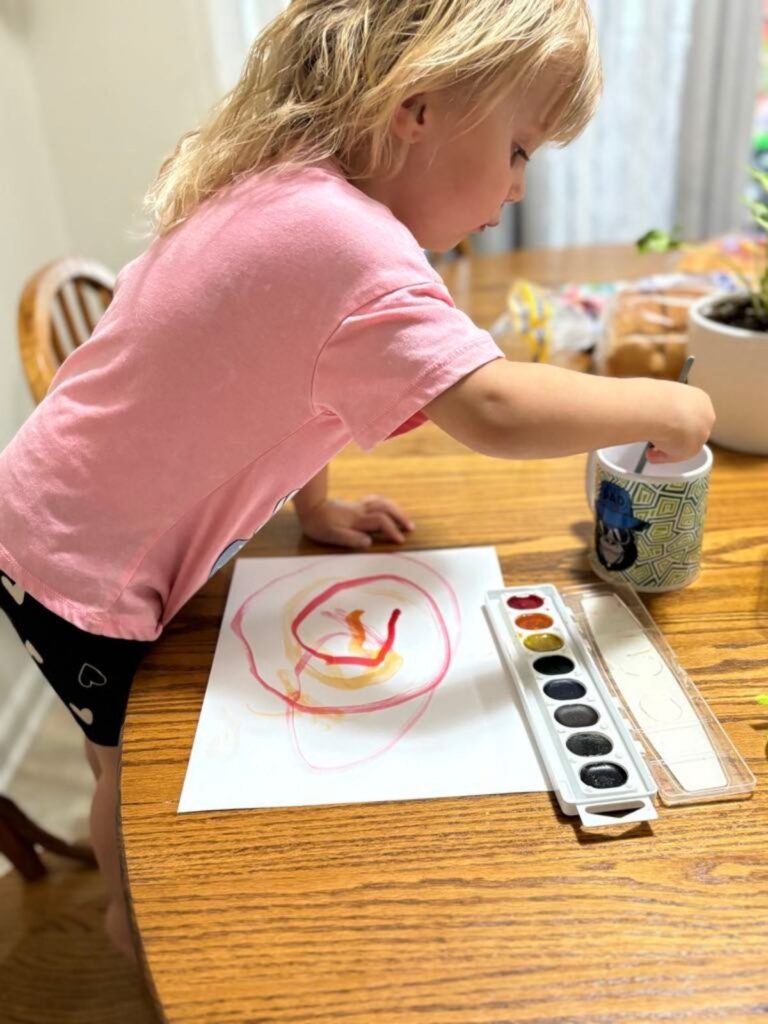 A young child in a pink shirt paints with watercolors at a wooden table, dipping a brush into a mug, their sticker earrings sparkling as they focus on the colorful drawing on paper in front of them.