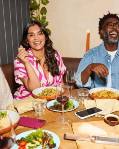 Three people sit at a table with plates of food, glasses of wine, and various dishes, smiling and enjoying a meal together indoors—perhaps celebrating their progress with Weight Watchers.