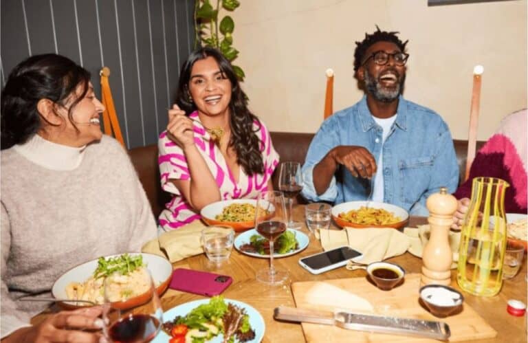Three people sit at a table with plates of food, glasses of wine, and various dishes, smiling and enjoying a meal together indoors—perhaps celebrating their progress with Weight Watchers.