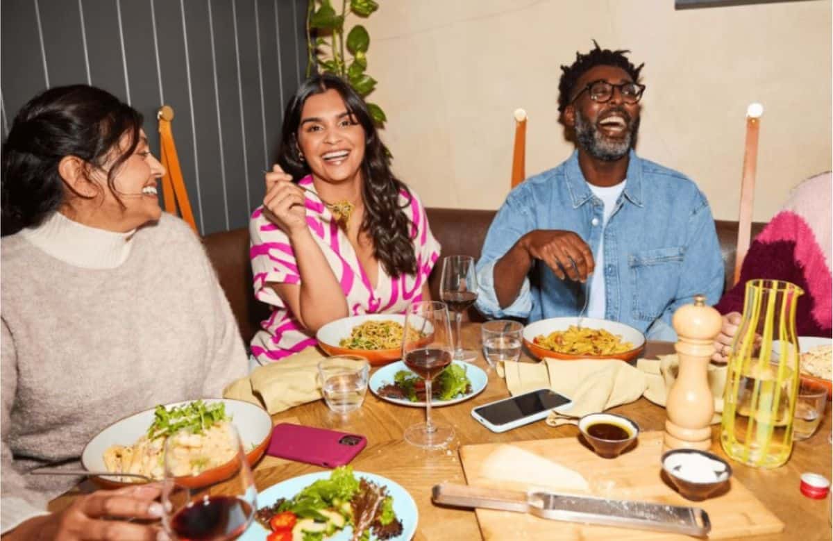 Three people sit at a table with plates of food, glasses of wine, and various dishes, smiling and enjoying a meal together indoors—perhaps celebrating their progress with Weight Watchers.