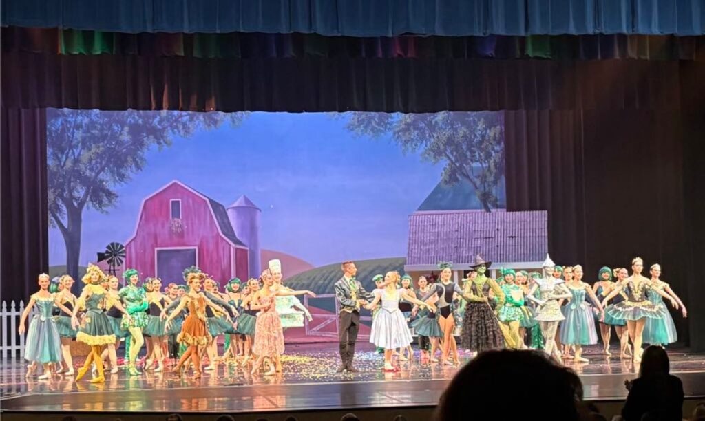 A group of dancers in colorful costumes perform on stage with a painted farm backdrop, including a barn and silo, capturing the energy of sweet spring under vibrant stage lights.