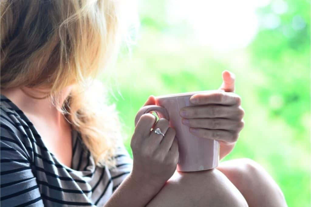 A person with blonde hair holds a pink mug and looks out a window, wearing a striped shirt and showcasing essential life skills like mindfulness. The background is blurred with greenery.