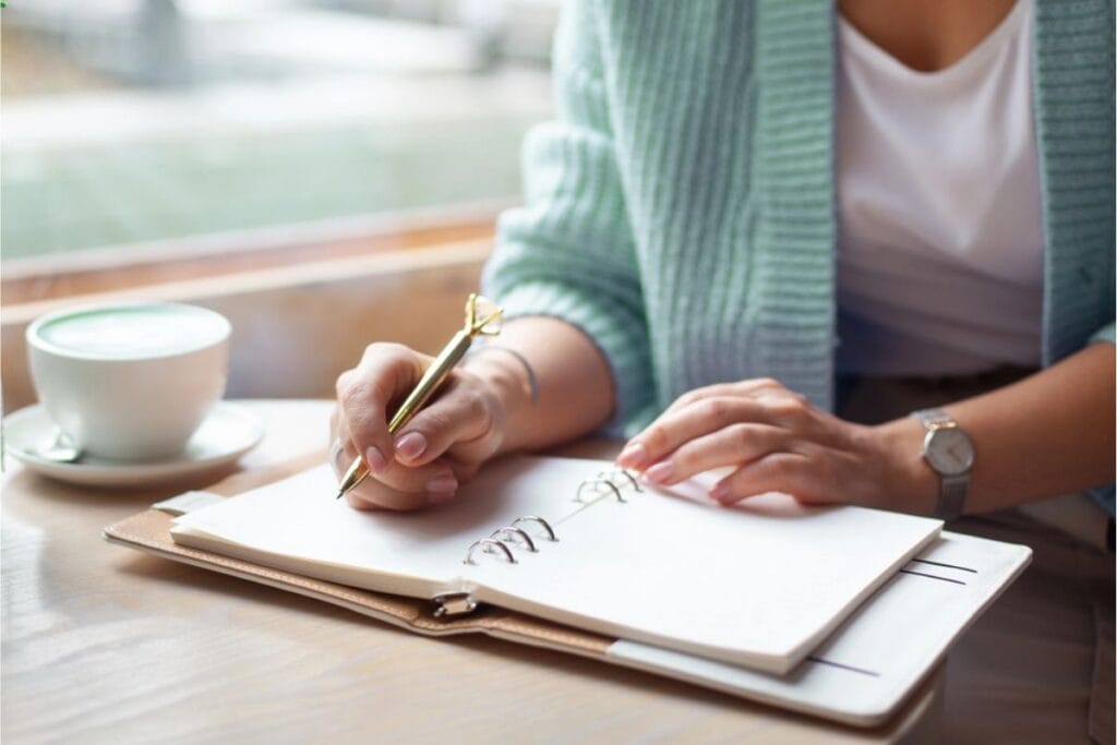 A person sits at a table writing in a notebook with a gold pen—practicing essential life skills—with a cup of coffee and eyeglasses nearby.