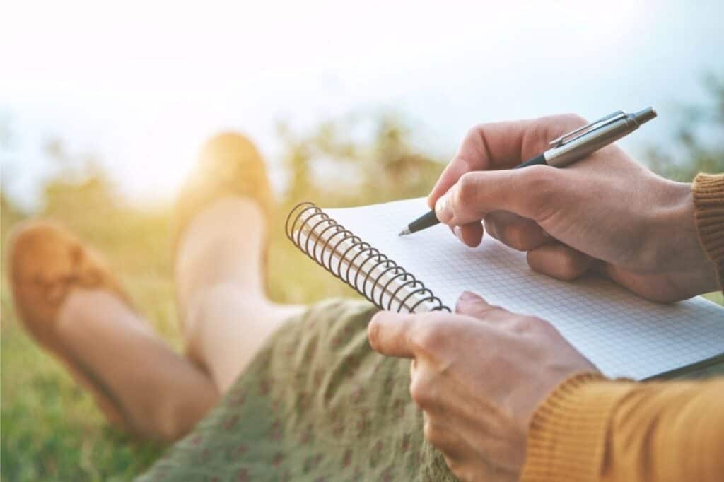 Person sitting on grass, holding a spiral notebook and pen, writing or drawing outdoors with sunlight in the background—practicing essential life skills like creativity and self-expression.