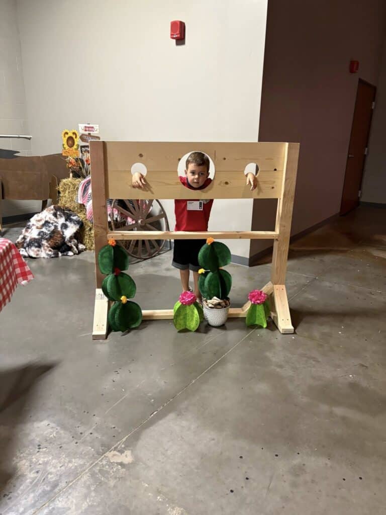 A young boy stands smiling with his head and hands through a wooden stocks prop, surrounded by artificial cactus decorations indoors.