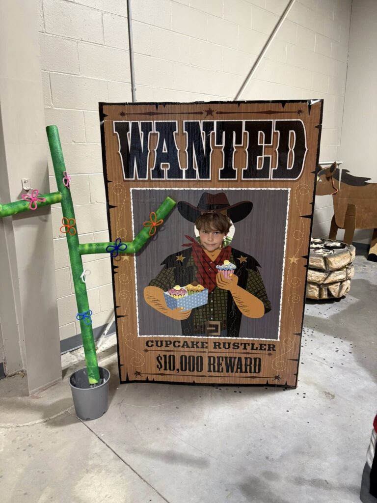 Person posing with their face in a "Wanted: Cupcake Rustler $10,000 Reward" photo standee, holding cupcakes. A cactus decoration is beside the standee indoors.