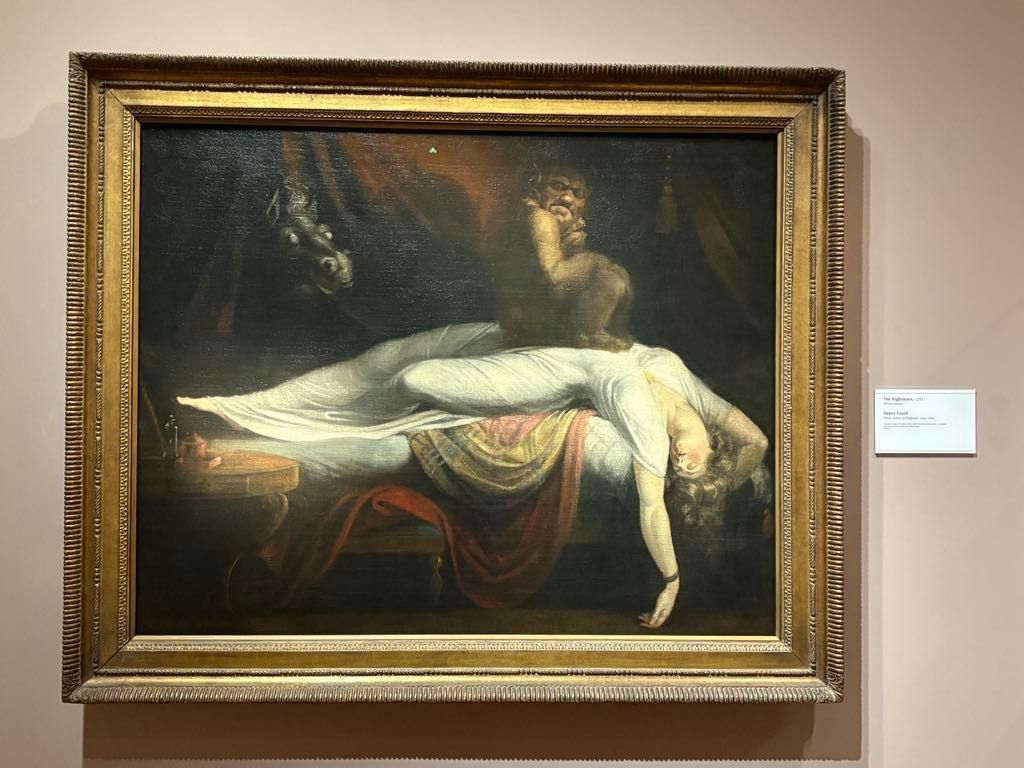 A painting depicts a woman in white draped across a bed while a dark, menacing figure crouches on her chest and a horse's head emerges from the shadows in the background.