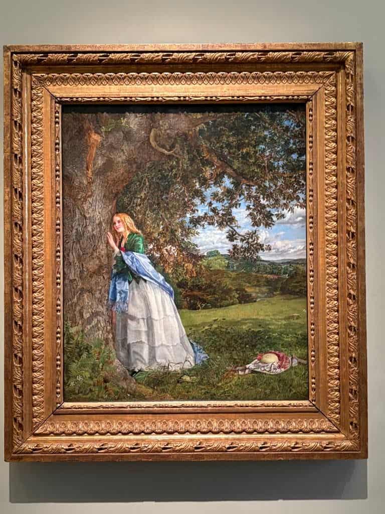 Framed painting of a woman in a blue and white dress standing by a large tree in a grassy landscape, with a hat placed on the ground nearby.
